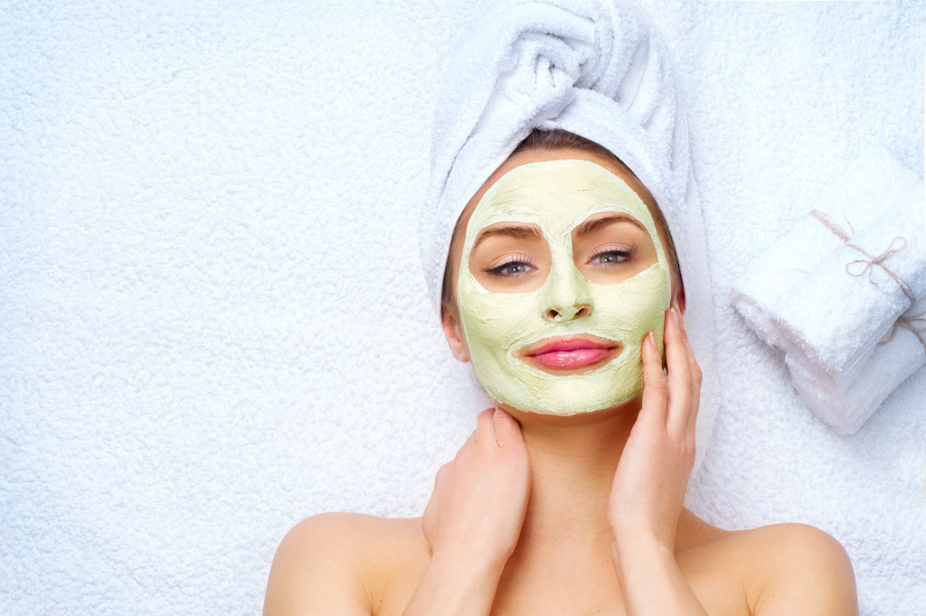 smiling woman touching her face with green homemade face mask lying in white towel